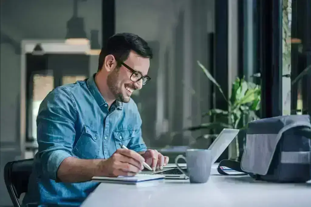 Man smiling at laptop screen while writing in notebook