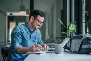 Man smiling at laptop screen while writing in notebook