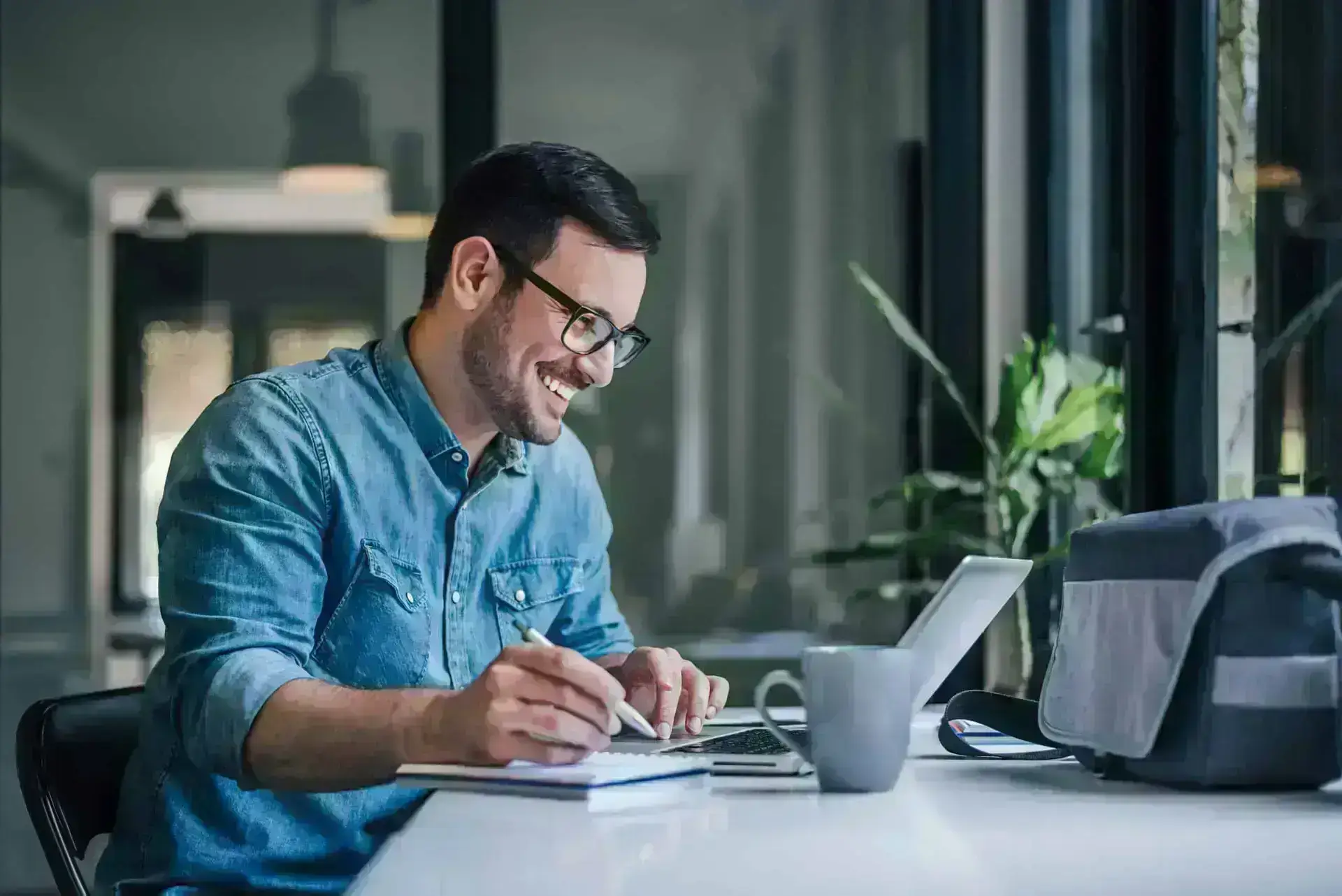 Man smiling at laptop screen while writing in notebook