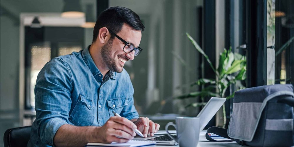 Man smiling at laptop screen while writing in notebook